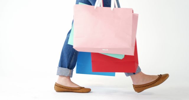 This image showcases a woman walking forward while holding colorful shopping bags. Her casual attire, including jeans rolled up at the cuffs and comfortable brown flat shoes, portrays a relaxed shopping experience. Ideal for advertisements, blog posts, and social media campaigns related to retail, sales, consumer behavior, and lifestyle. It can also be used to promote fashion, casual wear, and footwear.