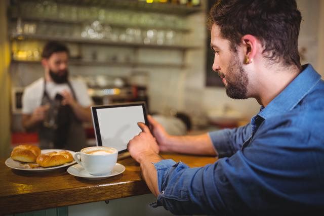 Man sitting at counter using digital tablet while enjoying coffee and croissants in a cafe. Barista in background preparing drinks. Ideal for illustrating modern lifestyle, technology use in everyday life, and casual dining settings.