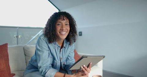 Image depicts an African American woman smiling and engaging with a tablet, exemplifying urban lifestyle and comfort. She sits in a contemporary loft, characterized by minimalist decor and warm accents like red pillows. This visual is perfect for promoting tech products, lifestyle blogs, interior design themes, or urban living articles. It's ideal for highlighting modern relaxation and convenience facilitated by technology in stylish living spaces.