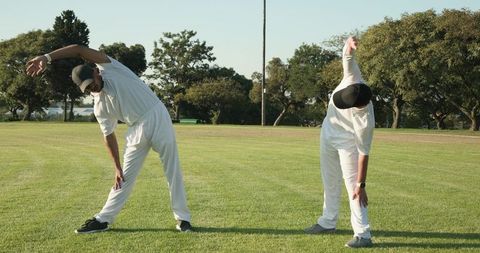 Cricket teammates stretching side bend on sunlit grassy field pre-match warmup