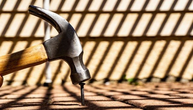 Hammer striking nail into brick pavers under striped sunlight for diy patio repair