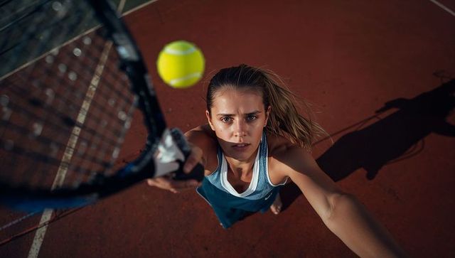 High-angle female tennis player reaching overhead and striking bright yellow volley on hardcourt