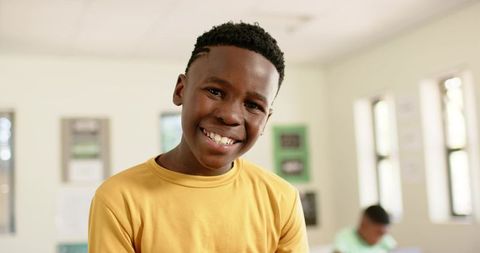 Smiling teen male leaning toward camera in sunlit classroom wearing mustard shirt