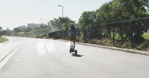 Young woman riding electric scooter on sunny urban road with backpack, shorts and cap