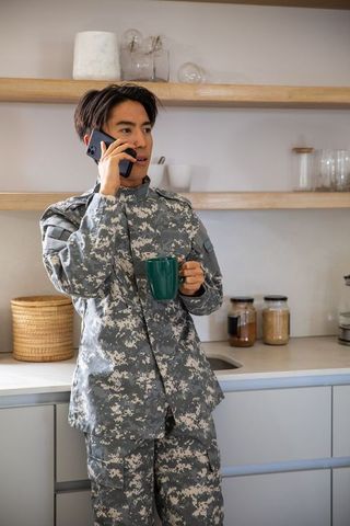Asian soldier talking on smartphone while holding mug in modern kitchen