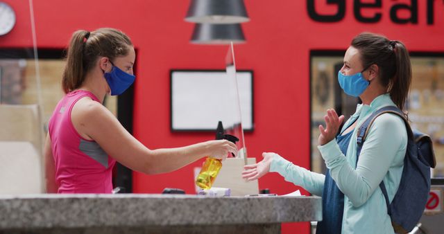 Woman in gym receiving hand sanitizer from staff member before working out. Both women wearing masks for safety. Can be used for health, fitness, safety, and hygiene topics during COVID-19 or other prevention efforts.