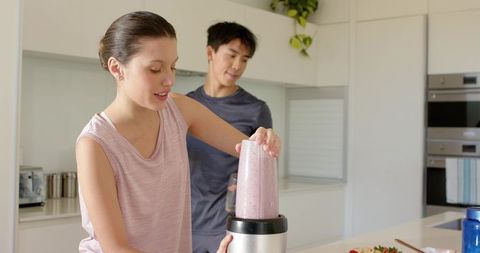 Couple preparing berry smoothie in bright modern kitchen, woman loading blender cup