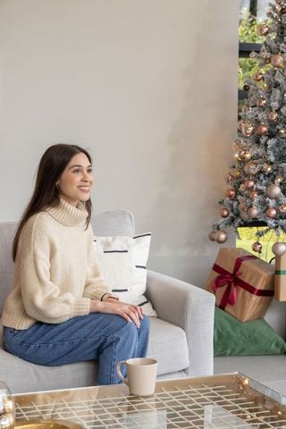 Woman wearing cream sweater sitting on sofa smiling beside decorated christmas tree with gifts