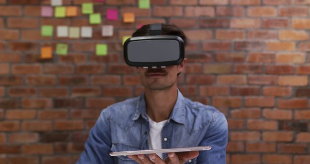 This visual depicts a person immersed in virtual reality while using a tablet in a contemporary office with a brick wall background. Suitable for promoting VR technology, tech startups, digital workspaces, or modern workspace environments.