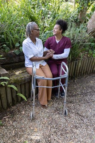 Maroon-scrub caregiver holding hands with senior african american woman on bench by walker