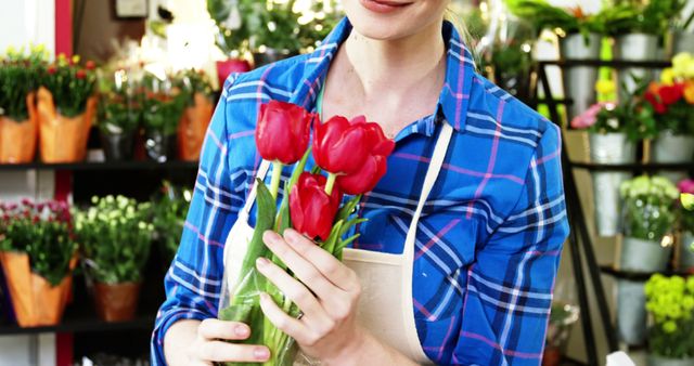 A female florist wearing a blue plaid shirt and apron holding a bouquet of fresh red tulips in a vibrant flower shop. Ideal for use in content related to small businesses, floral design, gardening, and retail environments. Perfect for blogs, advertisements, and marketing materials promoting floristry, florist services, or tutorials on flower arrangement.