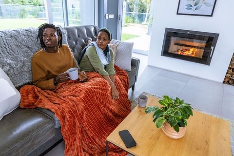 African american mother and teen daughter relaxing on couch by fireplace holding mugs