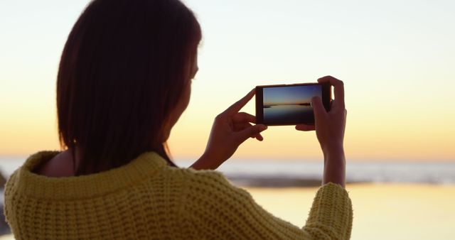 Woman standing on beach, capturing stunning sunset using smartphone. Midframe view focuses on device with vibrant sky and sun setting reflected in water. Useful for vacation, technology, calm nature themes.