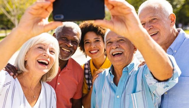 Seniors and adult friend taking joyful selfie sunlit park wearing colorful casual clothes