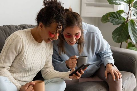 Friends sharing cozy self-care moment on couch wearing under-eye patches and holding mugs