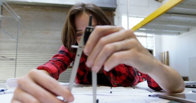 Young engineer or architect in a red plaid shirt using a compass to draft blueprints. This image is ideal for illustrating engineering, architecture, design, and creative work environments. It can be used for articles on the planning stages of construction projects, educational materials about engineering or architecture, or advertisements for engineering tools and resources.