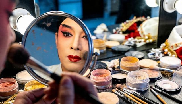Asian performer applying dramatic stage makeup at backstage vanity mirror, brushes