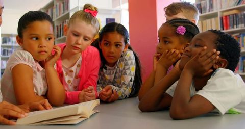 Diverse children leaning together reading open book in school library for literacy