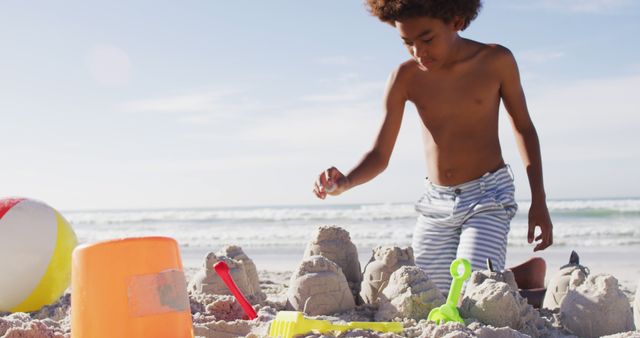A young boy is building sandcastles on a sandy beach under clear skies. He appears engrossed in his activity, using a pail, shovel, and various castle molds. In the background, the ocean waves are visible, indicating a seaside setting. The setting is perfect for use in summer vacation promotions, advertisements for children's toys, travel brochures, and kid-centered leisure activities.