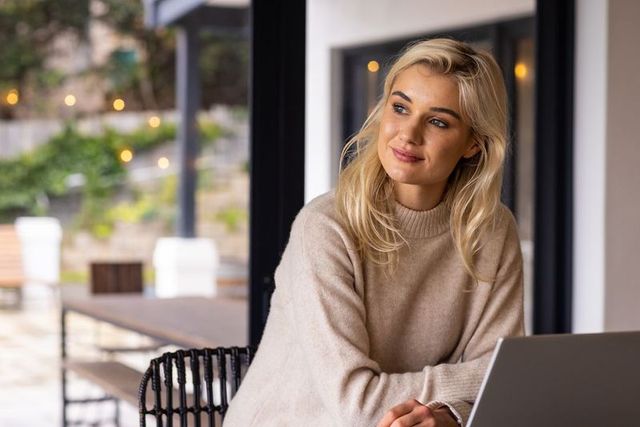Blonde woman working remotely on laptop from sunlit patio in beige sweater