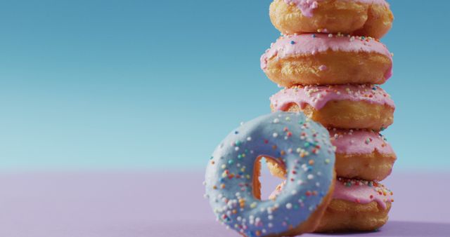 Brightly colored donuts with sprinkles are stacked against a blue and purple pastel background. Perfect for use in food blogs, bakery advertisements, dessert promotions, social media content, and culinary websites. The vibrant colors and appetizing presentation make it ideal for conveying themes of indulgence and delight.