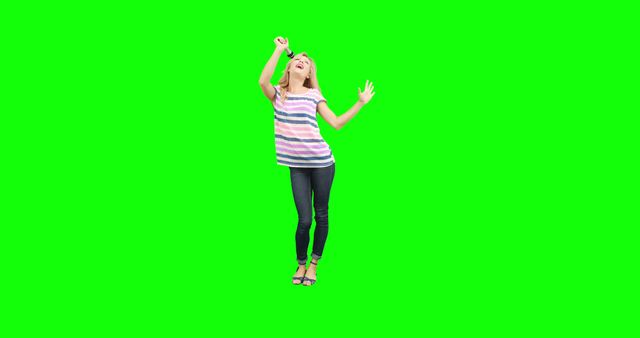 This image shows a joyful young woman in casual clothing, singing and dancing energetically against a green screen background. The vibrant green background makes it ideal for adding custom backdrops or special effects, perfect for marketing materials, advertisements, and promotional videos requiring dynamic and uplifting themes.