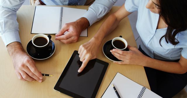 A Caucasian man and woman are engaged in a business meeting, reviewing information on a tablet, with copy space. Their professional collaboration is highlighted by the presence of coffee cups and notepads, suggesting a productive work session.