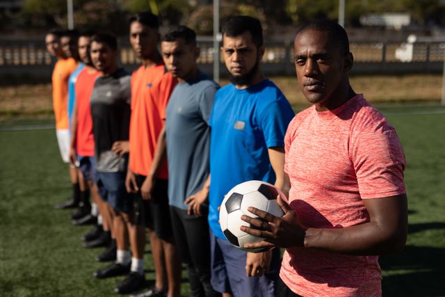 Portrait of multi ethnic team of male football players wearing sports clothes training at a sports field in the sun, standing in a row with one of players holding a ball.