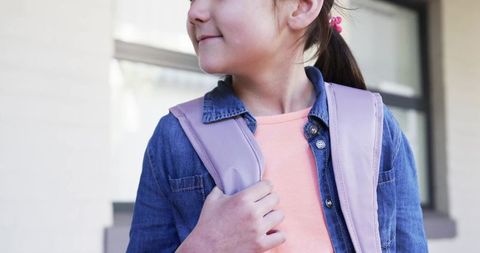 Schoolgirl carrying pastel pink backpack strap while standing outside wearing denim jacket