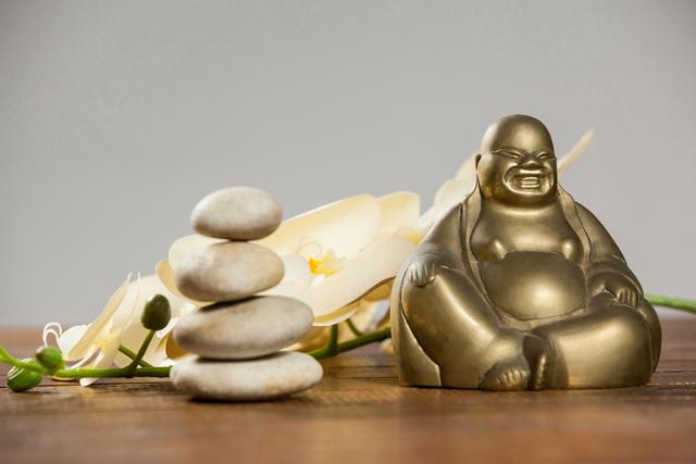 Laughing Buddha figurine sitting next to stacked pebbles and a white flower on a wooden table. Ideal for use in articles or advertisements related to meditation, relaxation, spirituality, wellness, and home decor. Perfect for illustrating concepts of balance, harmony, and tranquility.