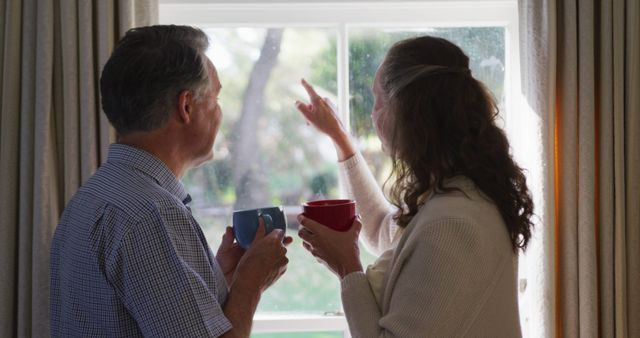 Rear view of senior caucasian couple looking out of window in living room, having coffee and talking. retirement lifestyle at home.