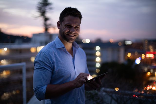 Man using a digital tablet while smiling at the camera during dusk. He is wearing a blue shirt and a watch, standing outdoors with a cityscape in the background. Ideal for use in business, technology, and professional lifestyle contexts.