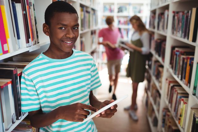 Schoolboy standing in library holding digital tablet, smiling at camera. Shelves filled with books in background, other students browsing. Ideal for educational content, technology in education, school promotions, and youth learning resources.