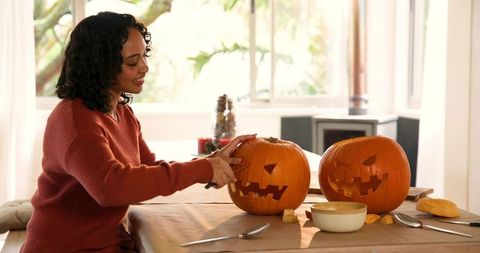 Woman carving jack-o'-lanterns in cozy autumn kitchen, preparing pumpkins for halloween