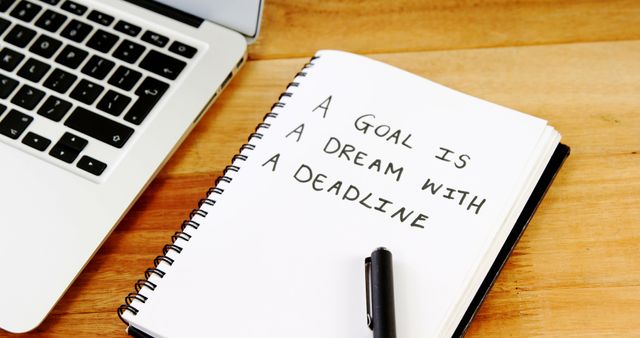 A notebook with the inspirational quote A Goal is a Dream with a Deadline lies next to a laptop and pen on a wooden desk, with copy space. It captures a motivational concept, often used to encourage productivity and goal setting.