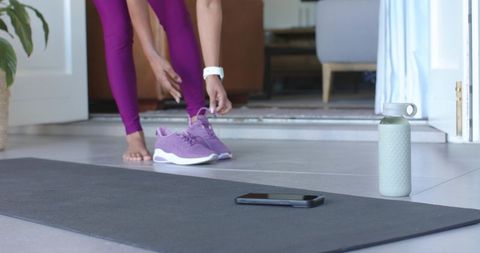 Indian woman putting on purple sneakers at home by yoga mat, smartphone and water bottle