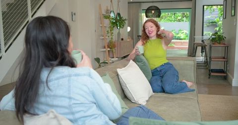Two diverse women chatting and sipping coffee on sectional sofa in bright open-plan living room