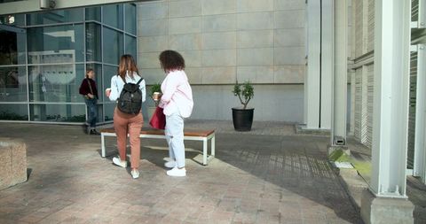 Diverse young women chatting over coffee near urban bench outside glass-fronted courtyard