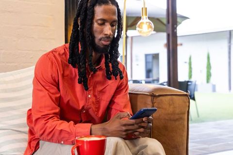 African american man using smartphone in modern cafe lounge with red mug and leather sofa