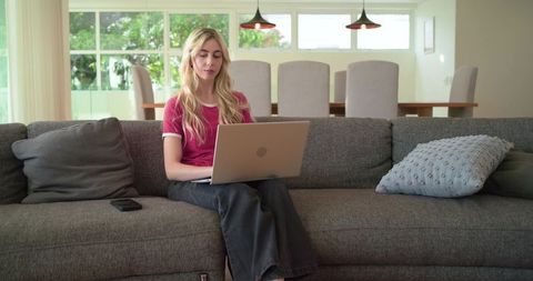 Blonde woman using laptop on gray sofa, remote work, home office, relaxed modern interior