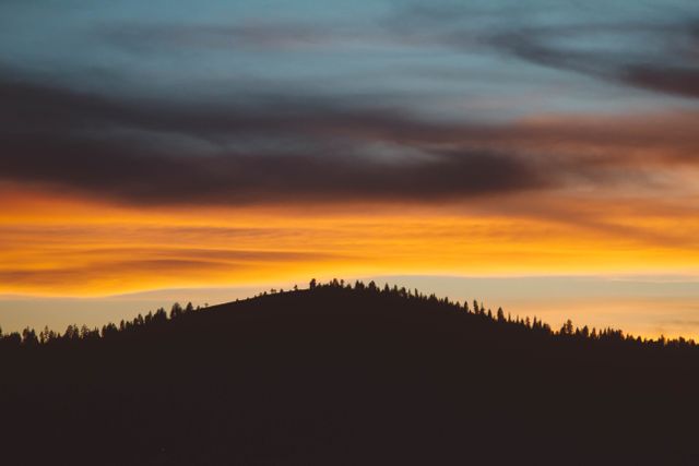 This image portrays a scenic and dramatic sunset with a forested hill as a silhouette. The sky exhibits vibrant colors transitioning from orange to deep blue, providing a tranquil and mesmerizing view. This image is ideal for use in travel magazines, nature blogs, or as a calming background image in presentations and websites dedicated to natural beauty and outdoor activities.