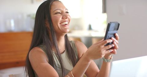 Young woman laughing while holding smartphone during video call in bright home workspace