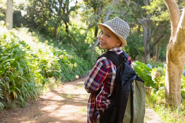Young boy wearing a plaid shirt and hat, carrying a backpack, smiling while looking back on a forest trail. Ideal for use in content related to outdoor activities, childhood adventures, nature exploration, hiking, and family outings.