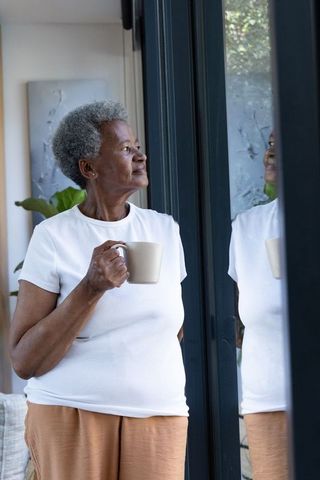 Senior african american woman standing by glass door holding mug and gazing outside