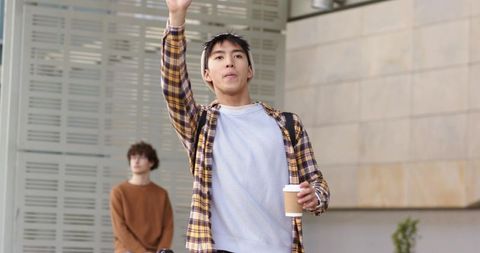 Young man raising hand while holding coffee and backpack outside modern urban building