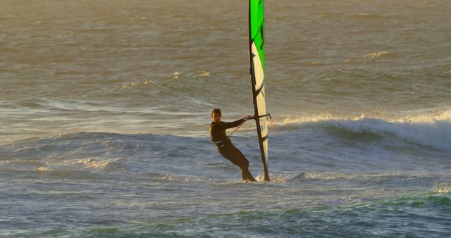 Windsurfer riding waves at sunset, ideal for promoting outdoor sports activities, adventure tours, coastal tourism, water sports equipment. Captures dynamic movement and athleticism against a picturesque ocean backdrop.