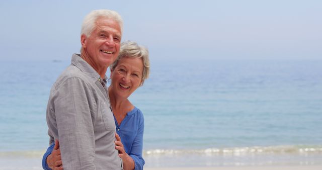 Senior couple hugging and smiling while standing on a sunny beach with the ocean in the background. Ideal for use in ads promoting retirement, senior living, travel, leisure activities, and happiness in later years. Suitable for lifestyle blogs, health and wellness content, and travel brochures.