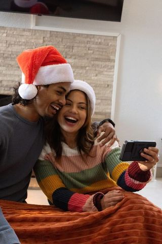Diverse couple wearing santa hats laughing while taking selfie on cozy living room couch