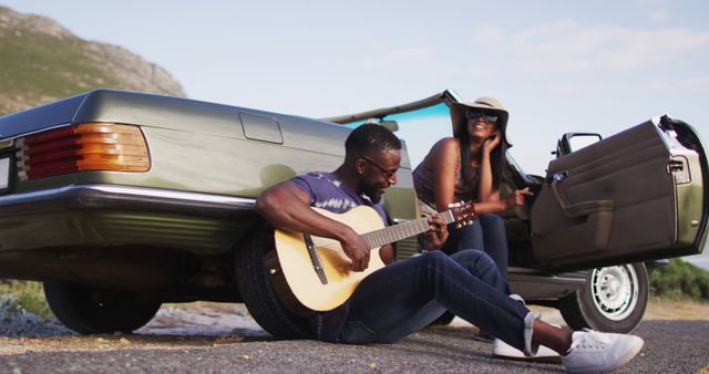 Couple enjoying road trip bonding while one is playing acoustic guitar and the other is sitting nearby, relaxing by vintage car beside mountains. Perfect for promoting experiences of relaxation and leisure during travel, romantic getaways, and summer adventures.