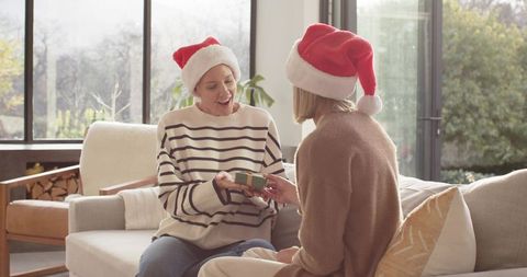 Mother and daughter exchanging christmas gift on sofa wearing santa hats cozy holiday moment